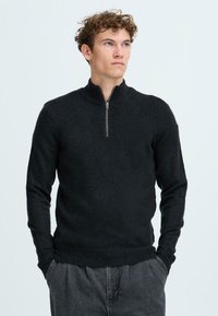 Schwarzer Pullover mit Reißverschluss am Hals und langen Ärmeln, aus weichem Strickmaterial gefertigt. Verfügt über einen gerippten Kragen und einen gerippten Saum und wird mit grauen, lässig geschnittenen Hosen kombiniert.