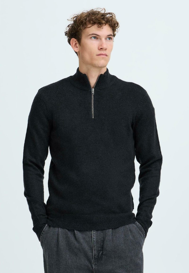 Black zip-neck sweater with long sleeves, made of soft knit material. Features a ribbed collar and hem, paired with grey relaxed-fit trousers.