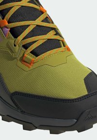 Chaussure de randonnée verte et noire avec tige synthétique texturée, lacets orange vif, et semelle extérieure noire robuste conçue pour l'adhérence.