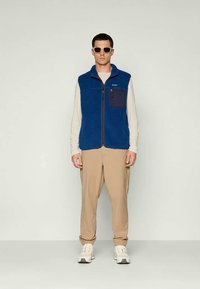 Modra flisasta jopica z zadrgo spredaj, ki ima temno modro žep. Nosi se nad svetlo dolgo rokavico in je v kompletu s bež hlačami ter belimi supergami.