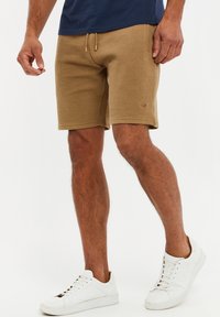 Homme portant un short marron texturé, une chemise bleu marine et des baskets blanches, debout devant un fond blanc.