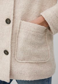Beige texturiertes Jackett mit großen Knöpfen und einer Hand, die teilweise in eine vordere aufgesetzte Tasche gesteckt ist, getragen über blauen Jeans.