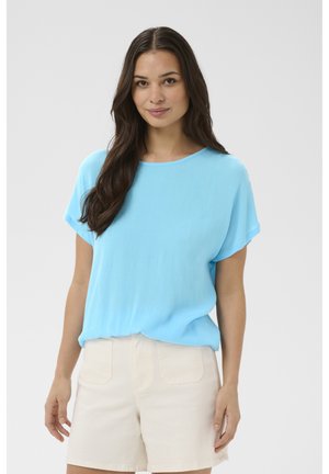 Kvinde med langt mørkt hår iført en lyseblå kortærmet top og beige shorts, stående foran en ensartet lys baggrund.