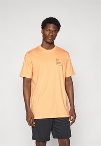 T-shirt en coton orange à manches courtes, avec un petit graphique texte noir sur la zone gauche de la poitrine. Assorti à un short de sport noir.