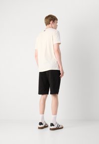 Light beige t-shirt with short sleeves, black shorts, and white sneakers with black accents. Model facing away from camera.