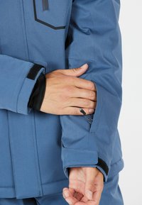 Person, die den Reißverschluss am Ärmel einer blauen Jacke mit schwarzem Innenbündchen und Klettverschluss am Handgelenk vor einem einfarbigen Hintergrund anpasst.