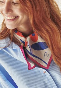 Barvna svilena ruta z abstraktnimi vzorci v rdeči, modri in krem barvi, zavezana okoli vratu. Nosi se čez svetlo modro srajco s črtami.