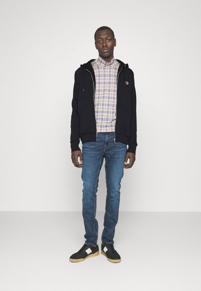 Svart zip-up hoodie, rutig skjorta i beige och blått, blå jeans och svarta skor med gummisulor, stående upprätt mot en enfärgad bakgrund.