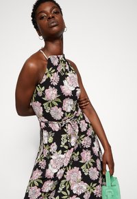 Vestido halter floral com tecido preto decorado com padrões florais em tons de rosa e verde, detalhe de fita na cintura, combinado com uma mala verde menta.