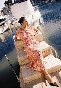 Femme en robe orange à motifs allongée sur des marches en pierre près d'une marina avec des bateaux blancs et de l'eau calme.