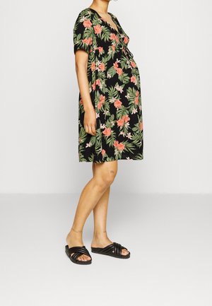Persona embarazada que lleva un vestido negro con flores hasta la rodilla y sandalias negras sin talón, de pie contra un fondo claro y liso.