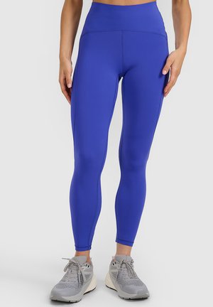 Vrouw die felblauwe hoge taille leggings en grijze hardloopschoenen draagt, staand op een effen lichtgrijze achtergrond.