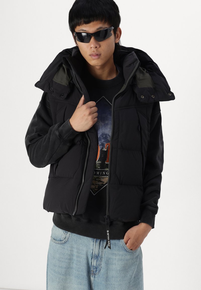 Chaleco tipo puff negro con capucha, con cierre de cremallera y botones a presión, superpuesto a una sudadera gráfica oscura, combinado con jeans de mezclilla azul claro.