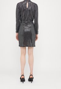 Robe mini noire à paillettes avec manches longues, design ajusté, détail de dos en trou de serrure et texture brillante. Associée à des sandales noires à talons ouverts.