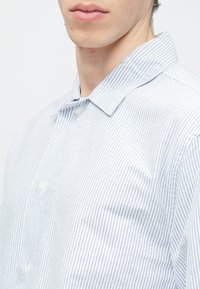 Artimos plano asmuo dėvi šviesiai mėlyną ir baltą dryžuotą marškinių viršutinę dalį su atverčia apykakle.