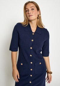 Marineblå strikket kjole med korte ærmer, stor krave, knaplukning foran, to forlommer og kontrastfarvede runde knapper. Glat tekstur.