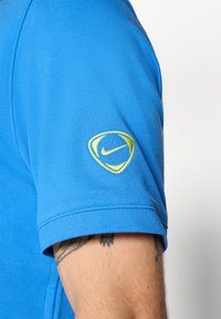 Nærbillede af en persons arm iført en blå kortærmet skjorte med et gult broderet Nike-logo på ærmet og synlige tatoveringer.