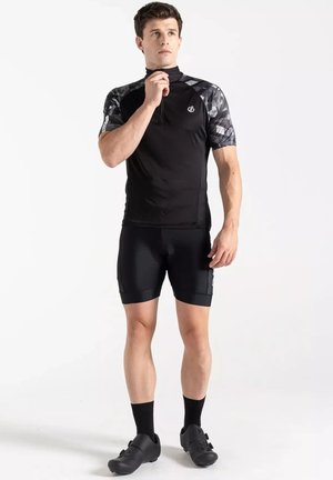 Jonge man die de kraag van een zwarte korte mouw fietstrui met patroon op de schouders, zwarte fietsbroek, sokken en schoenen verstelt.
