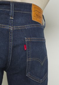 Vaqueros de mezclilla azul oscuro con costuras amarillas, una etiqueta de cuero marrón en la cintura y una pestaña de logotipo roja sobre el bolsillo trasero.