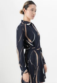 Femme portant une robe bleu marine avec des rayures abstraites beiges et blanches, à manches longues, avec un col et une ceinture assortie, se tenant contre un fond blanc.