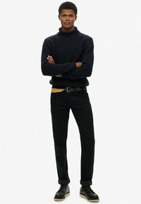 Dunkelblauer Strickrollkragenpullover, kombiniert mit einem gelben Hemd, schwarzen Jeans und schwarzen Stiefeln. Hat eine lässige Passform und aufgerollte Bündchen.