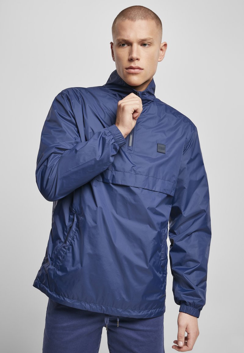 Jachetă de tip windbreaker de culoare albastru marin, cu guler înalt, fermoar pe jumătate în față, manșete elastice și buzunare laterale. Material neted, ușor.