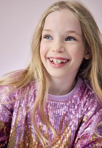 Glitrende pink paliettr sweater med horisontalt stribet design. Blød, kortærmet med en lys lilla krave. Lang, glat blondt hår.