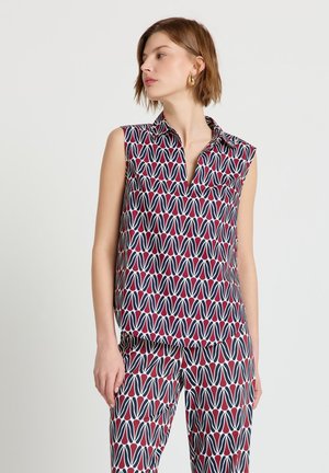 Donna con capelli castani corti che indossa un top senza maniche e pantaloni coordinati con un audace motivo geometrico rosso, blu navy e bianco, che guarda di lato.