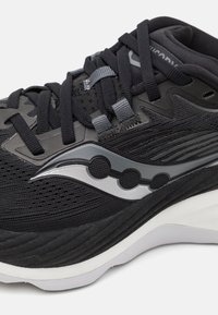 Svart träningsskor med en texturerad ovandel, grå accenter och en framträdande reflektiv logotyp. Sulan är vit med en lagerdesign.