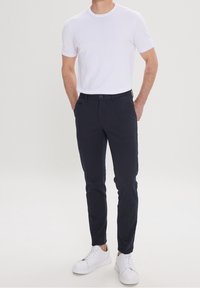 T-shirt blanc à manches courtes, ajusté, associé à un pantalon slim-fitt bleu marine. Le mannequin porte des baskets blanches. Fond uni, vue de face.