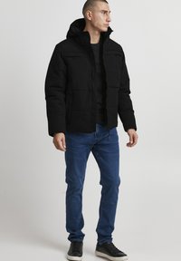 Sort polstret jakke med hætte, lynlåslukning og brystlommer. Kombination med blå jeans og sorte sneakers, stående mod en neutral baggrund.