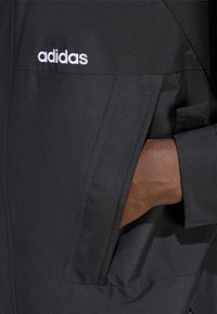Svart jacka med slät textur, med en ficka med dragkedja och en vit Adidas-logotyp. Handen är placerad i sidofickan.