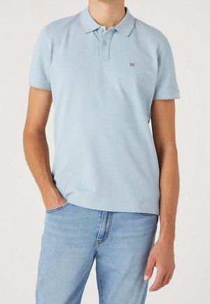 Jasnoniebieska koszulka polo z kołnierzykiem, krótkimi rękawami i subtelnym logo na piersi. Miękka tkanina o gładkiej fakturze. Noszona z jasnymi dżinsami.