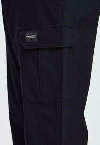 Black cargo pants featuring a side pocket with flap, sturdy fabric, straight cut, and visible stitching details. Brand tag on the pocket.