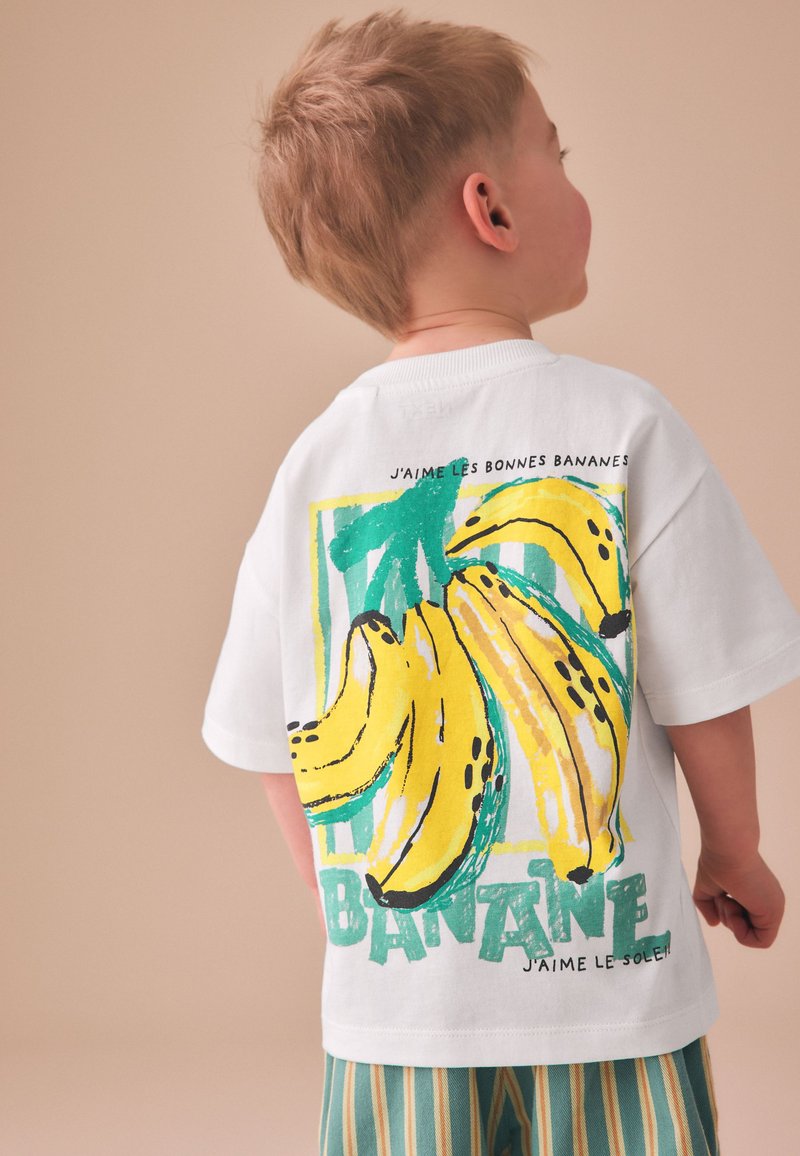 Niño con el pelo corto que lleva una camiseta blanca con una gran ilustración colorida de un plátano y texto en francés en la espalda.