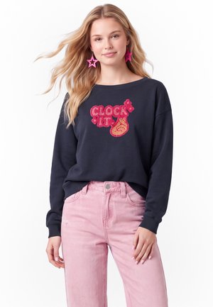 Ung kvinde med langt blond hår bærer en sort sweatshirt med teksten "CLOCK IT" og en lyserød flamme grafik, lyserøde jeans og store lyserøde stjerneøreringe.