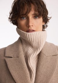 Płaszcz z wełny w odcieniu jasnobrązowym z grubym, prążkowanym, beżowym golfem. Zbliżenie podkreśla fakturę i detale projektu odzieży.