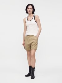 Femme portant un débardeur blanc avec un liseré noir, un short beige longueur genou et des bottines noires, debout devant un fond clair uni.