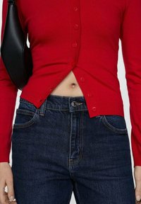 Rode aansluitende cardigan met knopen aan de voorkant, gecombineerd met donkerblauwe jeans. Inclusief een zwarte schoudertas. Effen kleuren en een gladde textuur.