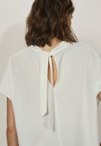 White top with a loose fit, short sleeves, keyhole back detail tied with a fabric ribbon, smooth texture.