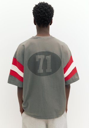 Persoană cu păr scurt și creț stă cu spatele, purtând un tricou gri cu mâneci cu dungi roșii și albe și un "71" negru pe spate.