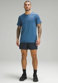 T-shirt de sport texturé bleu à manches courtes, associé à un short sombre et des chaussures de sport noires. Ajustement décontracté et design fonctionnel.