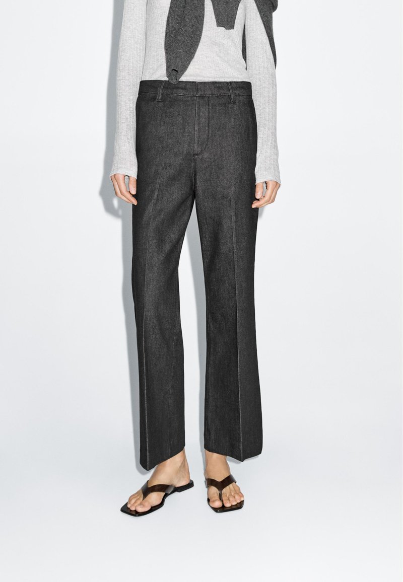 Donkergrijze broek met een rechte pijpdesign, met een soepel stoffen textuur en voorste plooidetails. Gepaard met een lichte trui en platte sandalen.