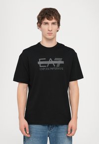 Black cotton t-shirt with a round neckline, featuring a gray graphic logo design "EA7 EMPORIO ARMANI" printed across the front. Short sleeves.