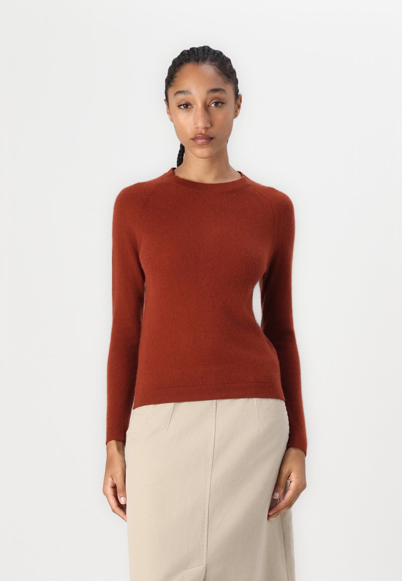 Rdzawoczerwony sweter z dzianiny, z długimi rękawami, okrągłym dekoltem i prążkowanym wykończeniem. Modelka łączy go z jasną spódnicą. Prosty i teksturalny wzór.