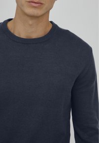 Marineblaues Rundhalspullover aus einem weichen Strickstoff, mit ribbeltem Halsausschnitt und langen Ärmeln. Glatte Textur mit minimalen Details.