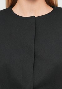 Μαύρο πλεκτό ύφασμα με στρογγυλή λαιμόκοψη και λεπτομέρεια στο μπροστινό κλείσιμο. Ομαλή υφή, μονόχρωμο, μίνιμαλ σχεδιασμός. Χωρίς σχέδια ή διακοσμήσεις.