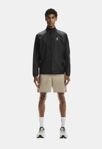 Melnā vieglā jaka ar augstu apkakli, priekšējo rāvējslēdzēju un logotipa detaļu. Valkāta virs bēšiem šortiem, kopā ar baltām zeķēm un sporta apaviem.