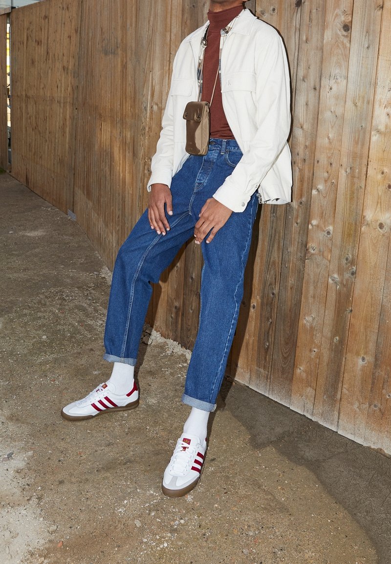 Vit kordjacka, brun polotröja, ljusblå denimjeans, vita sneakers med röda ränder, grå strumpor, stående mot en trävägg.