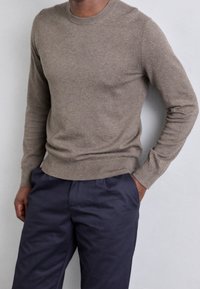 Bruine gebreide trui met lange mouwen en een geribbelde ronde hals, gecombineerd met donkerblauwe broek. De stof lijkt zacht en iets gestructureerd.